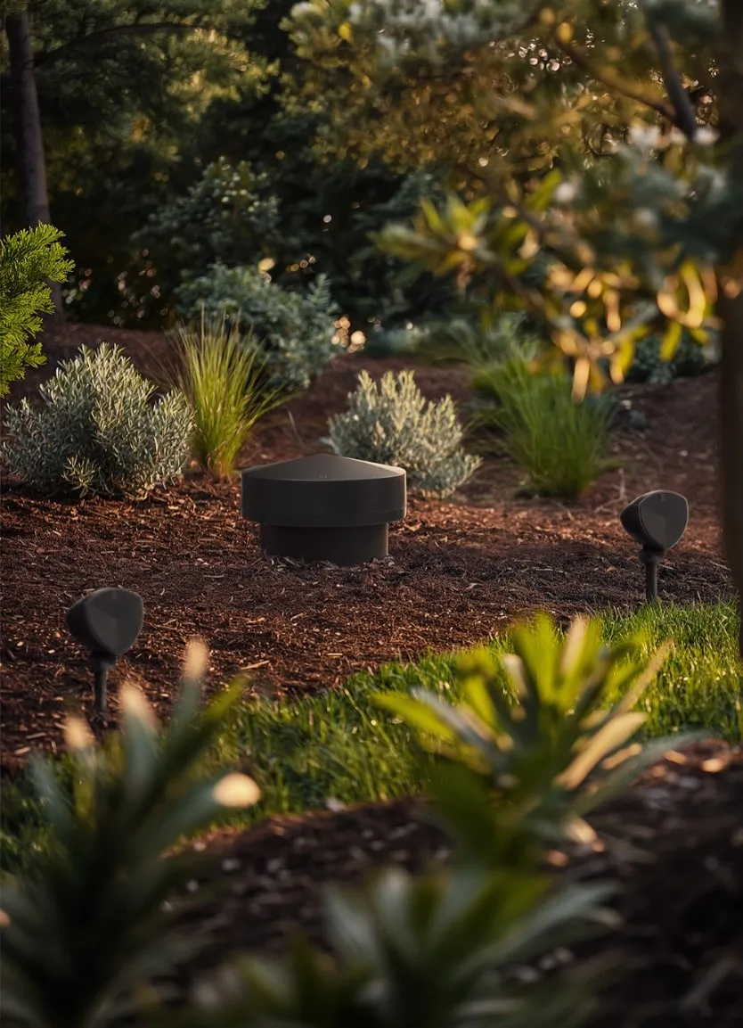 LS-80 subwoofer cap in a mulched garden bed at dusk with LS-400 satellites nearby